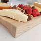 Wooden cutting board with assorted cheeses, fruits, and nuts on a white surface.
