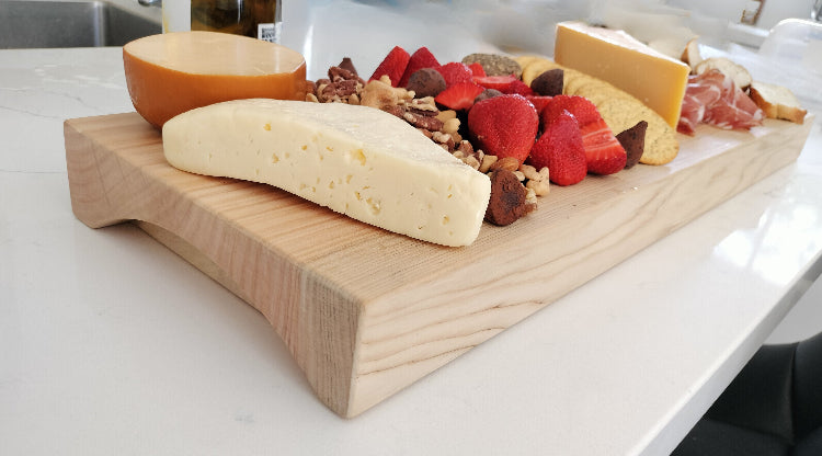 Wooden cutting board with assorted cheeses, fruits, and nuts on a white surface.